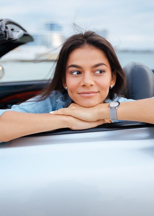 Smiling young woman sitting inside her convertible car on beach
